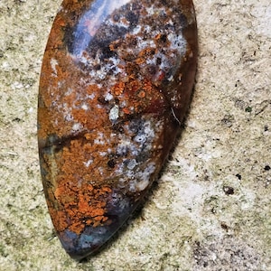 Puede incluir: Un cabujón de ágata pulido en forma de lágrima con un patrón marrón, naranja, blanco y gris. La piedra es translúcida y muestra una variedad de colores y texturas.