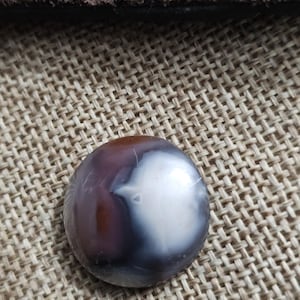 May include: A polished, round gemstone with a mix of colors. The stone features a blend of brown, gray, and white hues, with a smooth, reflective surface. The gemstone is set against a textured, woven background.