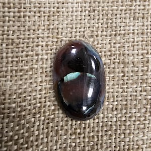 May include: A dark brown and black oval cabochon gemstone with iridescent green and blue accents. The gemstone is set on a burlap background.