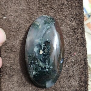 May include: An oval moss agate gemstone with a polished surface. The stone displays a mix of dark green, white, and translucent areas, forming a natural pattern. The agate is set against a brown background.