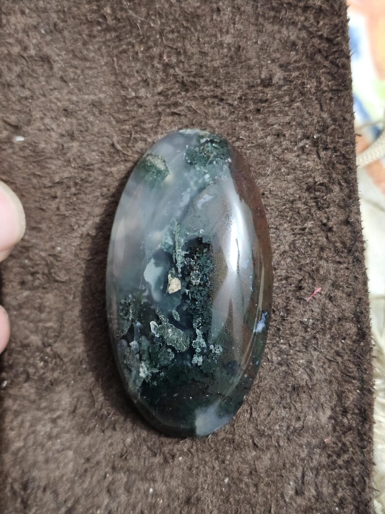 May include: An oval moss agate gemstone with a polished surface. The stone displays a mix of dark green, white, and translucent areas, forming a natural pattern. The agate is set against a brown background.