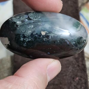 May include: An oval gemstone with a polished surface. The stone displays a mix of dark green, black, and white patterns, with a translucent quality. The gemstone is held between two fingers.