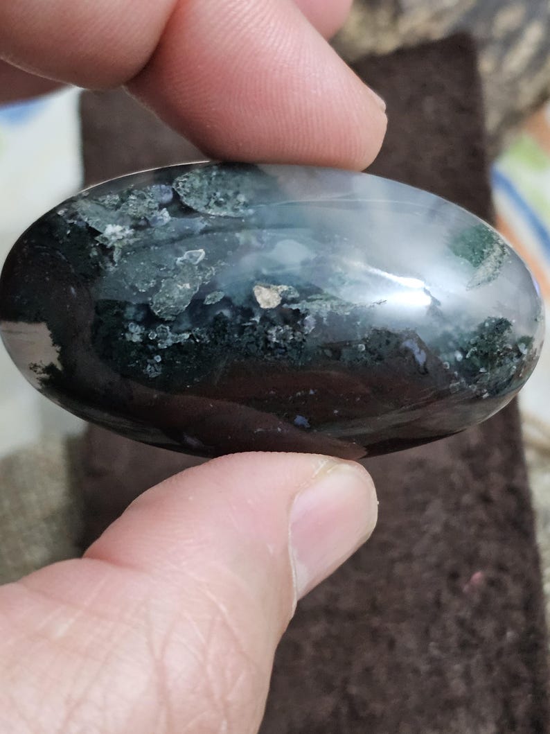 May include: An oval gemstone with a polished surface. The stone displays a mix of dark green, black, and white patterns, with a translucent quality. The gemstone is held between two fingers.