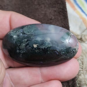 May include: A polished, oval gemstone displaying dark green, black, and purple tones. The stone has a smooth, glossy finish with lighter areas and small golden flecks. The gemstone is held in a hand.