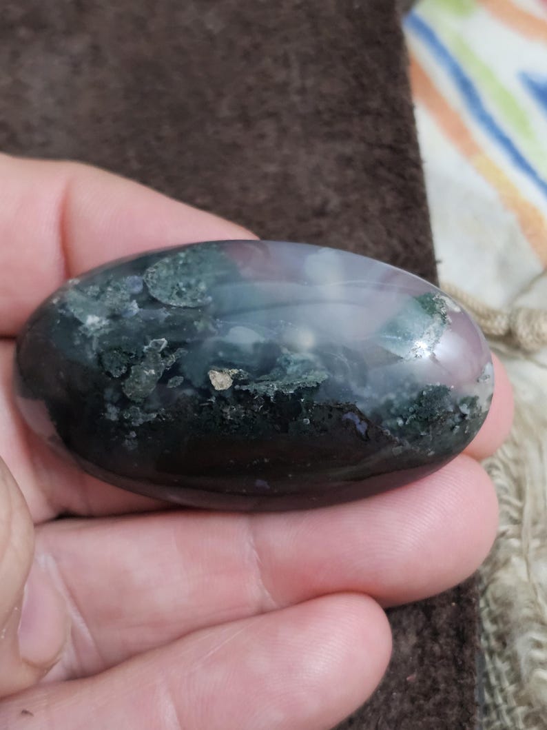 May include: A polished, oval gemstone displaying dark green, black, and purple tones. The stone has a smooth, glossy finish with lighter areas and small golden flecks. The gemstone is held in a hand.