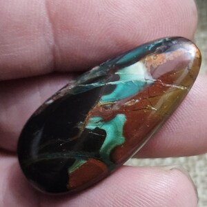 May include: A polished, oblong gemstone with a mix of colors. The stone features areas of black, brown, teal, and green. The colors blend and swirl together, creating a unique pattern. The stone is smooth and appears to be held in a hand.