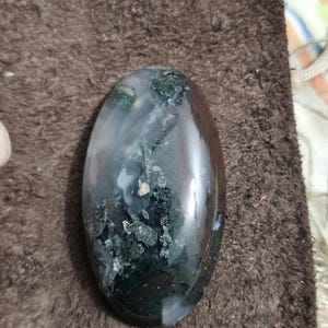 May include: A polished, oval gemstone displaying a mix of colours. The stone has areas of dark green, white, and brown, with a glossy finish. The gemstone is smooth and appears to be a natural stone.