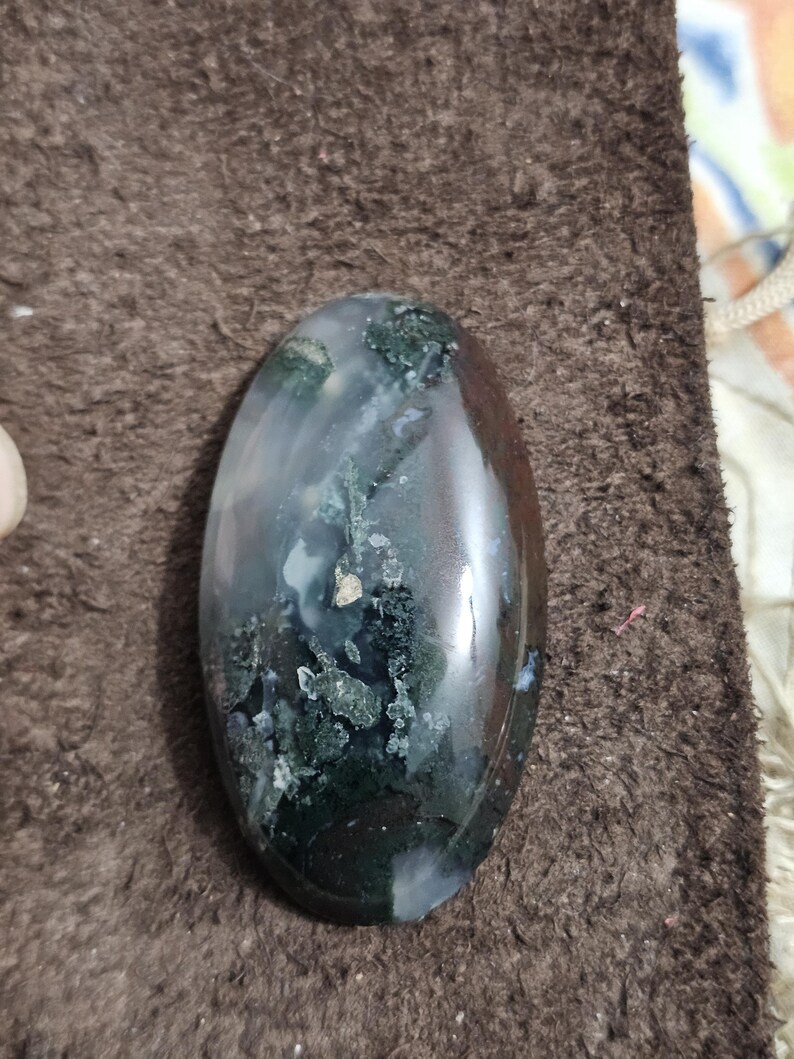 May include: A polished, oval gemstone displaying a mix of colours. The stone has areas of dark green, white, and brown, with a glossy finish. The gemstone is smooth and appears to be a natural stone.