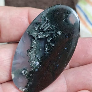 May include: An oval moss agate gemstone. The stone displays dark green, black, and white inclusions set against a translucent, pale purple and clear base. The polished surface reflects light, accentuating the natural patterns.