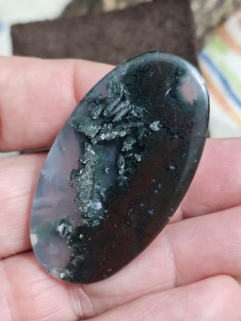 May include: An oval moss agate gemstone. The stone displays dark green, black, and white inclusions set against a translucent, pale purple and clear base. The polished surface reflects light, accentuating the natural patterns.