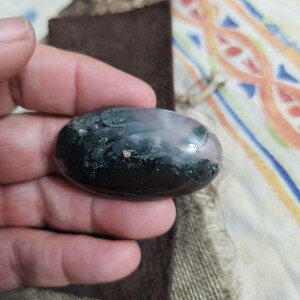 May include: An oval gemstone with a polished surface. The stone displays dark green, black, and light pink colours. The gemstone is held in a hand, with a brown suede-like material and a piece of wood in the background.