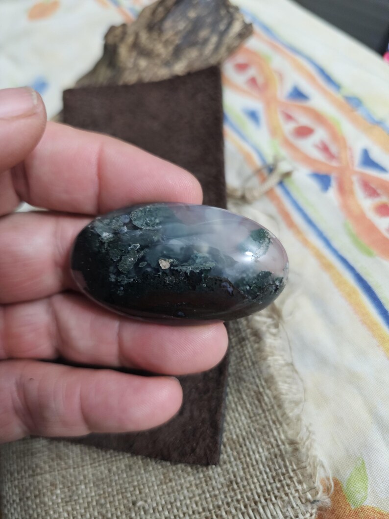 May include: An oval gemstone with a polished surface. The stone displays dark green, black, and light pink colours. The gemstone is held in a hand, with a brown suede-like material and a piece of wood in the background.