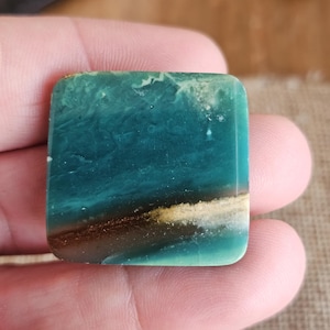 May include: A square, polished stone with a teal and brown color scheme. The stone has a marbled appearance with a band of brown and gold near the center. The edges are rounded, and the stone is held in a hand.
