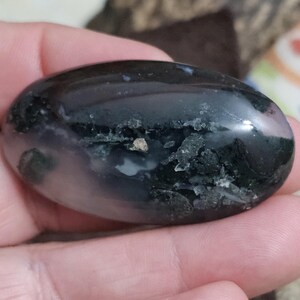May include: A polished, oval-shaped stone with a dark, glossy surface. The stone features areas of black, white, and green, with a small, light-coloured inclusion. The stone is held in a hand.