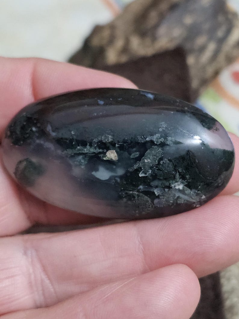 May include: A polished, oval-shaped stone with a dark, glossy surface. The stone features areas of black, white, and green, with a small, light-coloured inclusion. The stone is held in a hand.