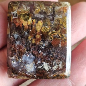 May include: A rectangular gemstone with a polished surface. The stone features a mix of colors, including brown, yellow, and white, with a marbled appearance. The stone is held in a hand.
