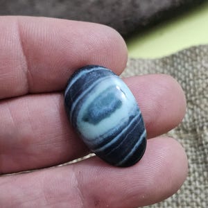 May include: An oval-shaped gemstone with a swirling pattern of dark blue, light blue, and white. The polished stone is held in a hand, showcasing its smooth surface and natural colors. The gemstone's design resembles an eye.