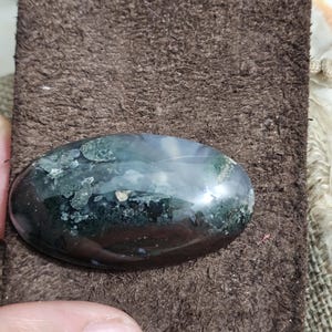 May include: An oval gemstone with a polished surface. The stone has dark green, black, and white colours. The gemstone is set against a brown, textured background.