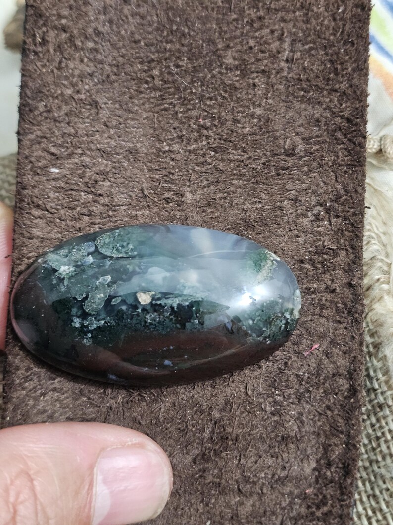May include: An oval gemstone with a polished surface. The stone has dark green, black, and white colours. The gemstone is set against a brown, textured background.