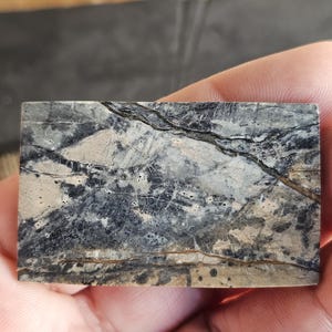 Könnte beinhalten: Ein rechteckiger Stein mit Marmormuster. Der Stein weist eine Mischung aus grauen, schwarzen und beigen Farben mit natürlichen Adern und Rissen auf. Der Stein wird in der Hand einer Person gehalten.
