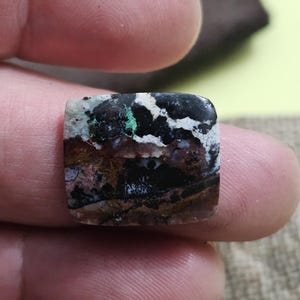 May include: A square-shaped gemstone with a unique pattern of black, white, green, brown, and purple hues. The stone is held between two fingers, showcasing its polished surface and intricate color variations. The gemstone is likely used for jewelry or decorative purposes.