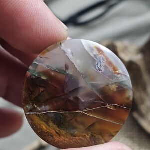 May include: A round, polished gemstone with a mosaic of colors including brown, orange, white, and green. The stone has a cracked, natural appearance, with visible fissures and variations in color. The gemstone is held between two fingers.