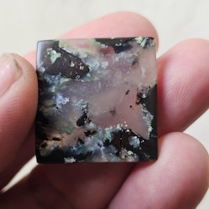 May include: Square gemstone with a mix of colors. The stone features a blend of black, green, and pink hues, creating a unique pattern. The edges are smooth, and the overall shape is a perfect square. The gemstone is held between fingers.