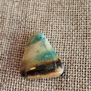 May include: A triangular, polished stone with a blend of colors. The top section is a light teal, transitioning to tan and then a dark brown band at the bottom. The stone has a glossy finish and is set against a textured, woven background.