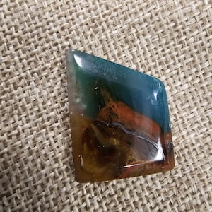 May include: A polished gemstone with a mix of colors, including teal, brown, and orange. The stone has a unique, irregular shape and a glossy finish. The gemstone is set against a neutral woven background.