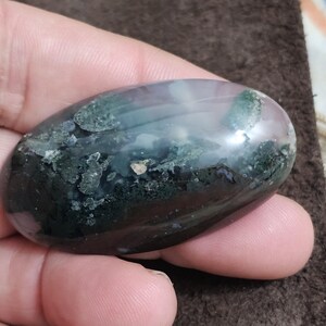May include: A polished, oval-shaped stone with a blend of colours. It displays areas of dark green, light blue, and a touch of pink. The stone is held in a hand, highlighting its smooth, rounded shape. This is a natural gemstone.