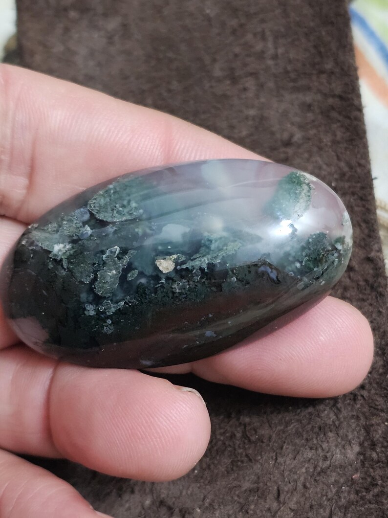May include: A polished, oval-shaped stone with a blend of colours. It displays areas of dark green, light blue, and a touch of pink. The stone is held in a hand, highlighting its smooth, rounded shape. This is a natural gemstone.