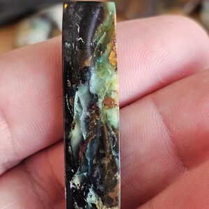 May include: A rectangular gemstone with a polished surface. The stone features a mix of colors, including black, green, brown, and hints of blue. The pattern is organic and flowing, with a marbled appearance. The gemstone is held in a hand.
