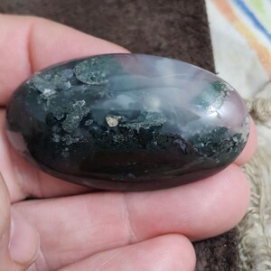 May include: An oval gemstone with a polished surface. The stone displays a mix of colours, including dark green, black, and hints of pink and white. The gemstone is held in a hand, showcasing its smooth texture and natural patterns.