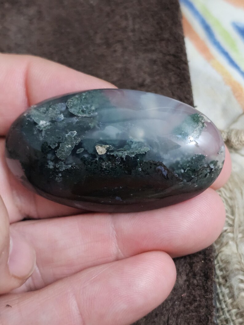 May include: An oval gemstone with a polished surface. The stone displays a mix of colours, including dark green, black, and hints of pink and white. The gemstone is held in a hand, showcasing its smooth texture and natural patterns.