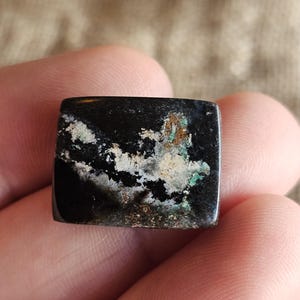 May include: A square, polished gemstone with a black base color and white, green, and brown mineral inclusions. The stone is held in a hand, showcasing its natural patterns and textures. The gemstone is likely used for jewelry or decorative purposes.