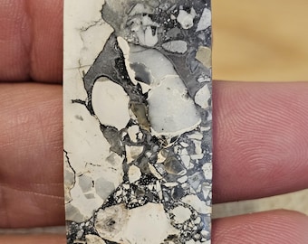 May include: A white and black speckled stone cabochon with a rectangular shape. The stone has a unique pattern of black and white swirls and speckles.