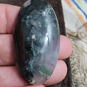 May include: An oval moss agate gemstone. The polished stone displays dark green, white, and translucent sections. The stone is held in a hand, highlighting its smooth, reflective surface. The agate has a natural, organic appearance.