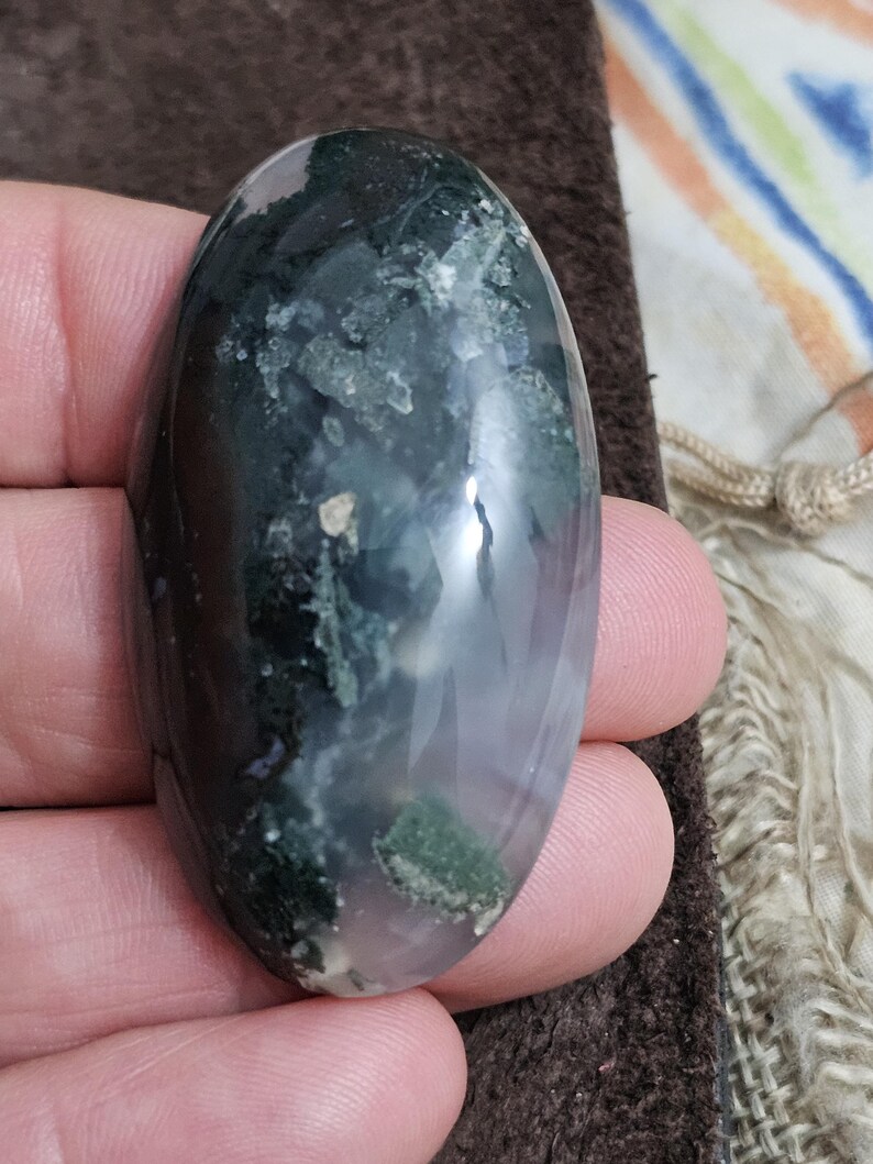 May include: An oval moss agate gemstone. The polished stone displays dark green, white, and translucent sections. The stone is held in a hand, highlighting its smooth, reflective surface. The agate has a natural, organic appearance.