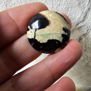 May include: A round, black and white cabochon with green and brown accents. The cabochon is smooth and polished.