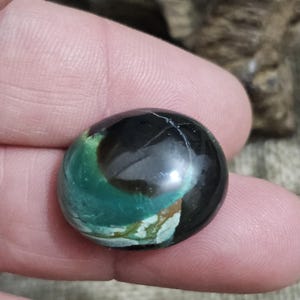 May include: A polished, round gemstone with a swirling pattern of black, teal, and brown. The stone has a glossy finish and appears to be held in a person's hand. The gemstone's unique color combination and shape make it a striking piece.