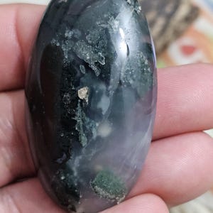 May include: An oval moss agate gemstone, displaying a mix of dark green, black, and translucent sections. The stone has a polished surface with visible inclusions and patterns. The colours shift from dark to light, creating a unique visual effect.