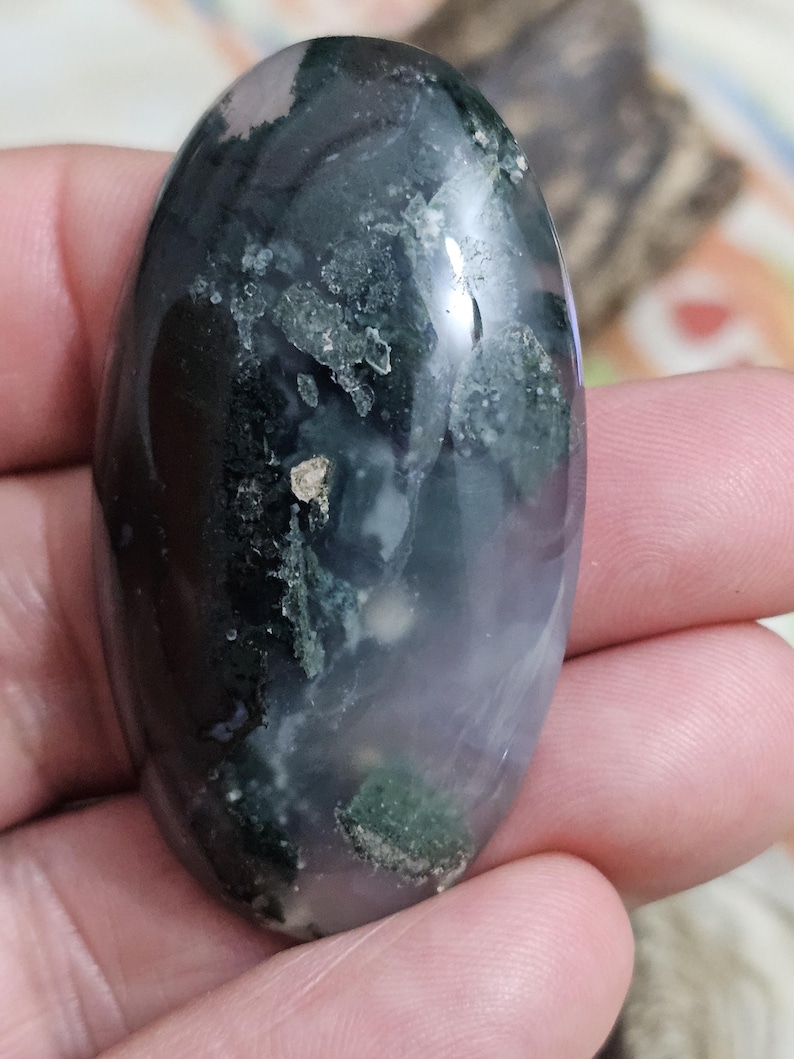May include: An oval moss agate gemstone, displaying a mix of dark green, black, and translucent sections. The stone has a polished surface with visible inclusions and patterns. The colours shift from dark to light, creating a unique visual effect.