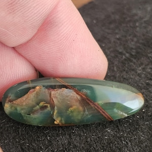 May include: An oval-shaped gemstone with a polished surface. The stone features a blend of green, brown, and tan colors, with a prominent brown streak running across its surface. The gemstone is held between fingers.