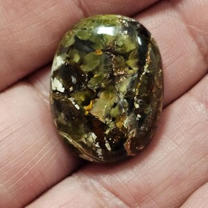May include: A close-up of a green, brown, and white oval cabochon gemstone with a copper-like sheen. The stone has a unique, marbled pattern.