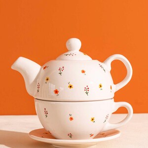 Może przedstawiać: Biały ceramiczny zestaw do herbaty z czajniczkiem, filiżanką i spodkiem. Zestaw jest ozdobiony małymi, kolorowymi wzorami kwiatowymi w kolorze czerwonym, żółtym i pomarańczowym. Czajniczek ma pokrywkę i zakrzywioną wylewkę. Filiżanka znajduje się w czajniczku.