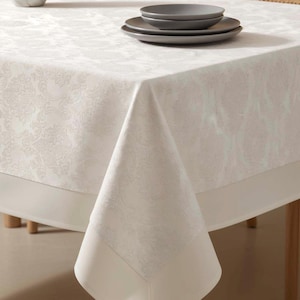 May include: A white tablecloth with a subtle floral pattern and a solid white border. Two gray plates are stacked on top of the tablecloth. The table is set with a vase and chairs in the background.