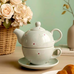 Puede incluir: Juego de té individual de cerámica azul claro con tetera, taza y platillo. El juego está decorado con pequeños puntos rojos y motivos florales. Una cesta de rosas y un cruasán están en el fondo.
