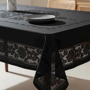 May include: A black tablecloth with a floral lace border and matching design in the center. Two gray plates are stacked on the table. A white and gray marble vase is also on the table.
