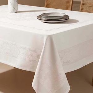 Könnte beinhalten: Eine weiße Tischdecke mit zartem floralem Spitzenrand. Zwei graue Keramikteller sind auf dem Tisch gestapelt. Eine weiße Marmorvase mit einer grünen Pflanze befindet sich im Hintergrund. Der Tisch ist für eine Mahlzeit gedeckt.