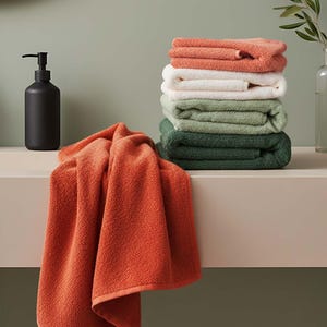 May include: A stack of folded towels in shades of orange, white, green, and dark green. A single orange towel is draped over the edge of a shelf. A black soap dispenser is to the left. The towels are made of a soft, textured material.